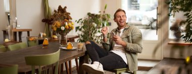 Kafedeki mutlu genç adam, cappuccino içiyor, kulaklıkla komik podcast dinliyor, akıllı telefon tutuyor. Yakışıklı erkek kafe ziyaretçisi, favori şarkısını cep telefonu müzik uygulamasında açıyor..