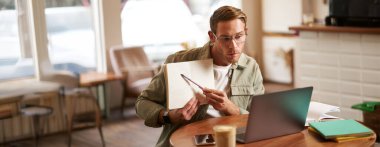 Gözlüklü yakışıklı bir adamın portresi, boş bir kafede çalışıyor, defterinde örnekler gösteriyor, ders veriyor, bilgisayarını öğrenciyle çevrimiçi görüşmek için kullanıyor..