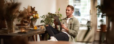 Kafede tek başına oturup dijital tablet okuyarak video izleyip, kablosuz kulaklıkla müzik dinleyen genç bir adamın portresi. Yaşam tarzı ve insan kavramı.