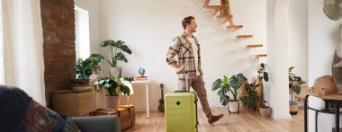 Evinde bavuluyla yürüyen, tatilde oda kiralayan, gülümseyen ve mutlu görünen yakışıklı genç bir adamın portresi. Seyahat ve turizm konsepti