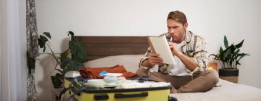 Kızgın bir adamın portresi, bavul hazırlıyor, tatil için kontrol listesiyle defterde hayal kırıklığına uğramış görünüyor, tatile çıkmaya hazırlanıyor. Turist ve insan konsepti