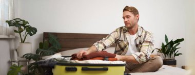 Giysilerini bavula dolduran genç bir adamın portresi, bavulla yatakta oturan, tatile çıkan, bir yolculuğa hazırlanan. Yaşam tarzı ve turizm konsepti