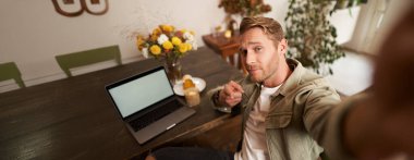 Cep telefonuyla selfie çeken yakışıklı ve kendine güveni tam bir genç. Kamerayı işaret ediyor. Kafede dizüstü bilgisayarla oturuyor..