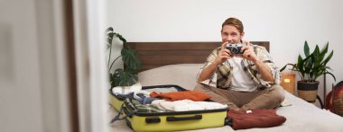 Turist portresi, bavuluyla yatakta oturan, kamera taşıyan, bavulunu kıyafetlerle dolduran, tatile çıkan, yaz tatiline hazırlanan, yurt dışına seyahat eden bir adam..