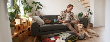 Köpekli bir adam bavulun yanında oturuyor, elinde defter var, tatile çıkarılacak eşyaların listesini kontrol ediyor, tatil gezisi planlıyor..