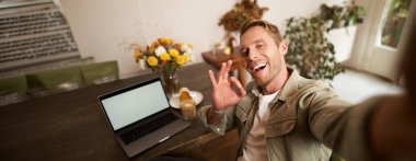 Yakışıklı, gülümseyen, selfie çeken, iyi el işareti gösteren, cep telefonuyla fotoğraf çeken, laptopuyla kafede oturan genç bir adam..