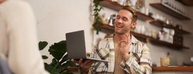 Genç gülümseyen adamın portresi, iş arkadaşı kafeden çevrimiçi toplantıya katılıyor, bilgisayardaki video sohbetleri, tamam, el imzası, kahve içme ve uzaktan çalışma..