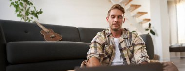 Bilgisayarı kullanan genç adamın portresini kapat, bilgisayara bak, yerde cihazıyla otur, web sitelerine bak.