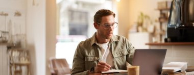 Modern bir adamın gözlüklü portresi, boş bir kafede dizüstü bilgisayarla oturuyor, not defterine notlar yazıyor, internetten okuyor, uzaktan öğreniyor, web kursundan geçiyor..