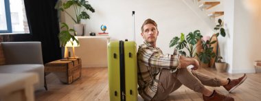 Erkek modelin bavullu portresi, yerde oturuyor, uçak biletleri ve pasaport tutuyor, yurtdışına gidiyor, havaalanına gitmek için taksi bekliyor..