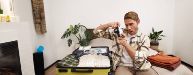 Genç bir adamın görüntüsü, turist kamerasını kontrol ediyor, açık bavulla yatakta oturuyor, fotoğraf çekiyor, giysilerini çıkarıyor, tatile çıkıyor. Seyahat, insan ve turizm konsepti