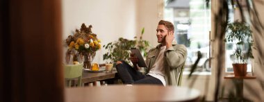 Yakışıklı, gülümseyen genç bir adamın portresi, kahve dükkanında oturuyor, dijital tablet kullanıyor, kafeden biriyle görüntülü sohbet ediyor, kablosuz kulaklık takıyor..