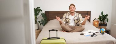 Yaz tatili için bavulunu topladıktan sonra yatakta meditasyon yapan genç bir adamın portresi, bacak bacak bacak üstüne atmış, nirvana pozu veren, rahatlatıcı, bir şeyler umut eden. Turizm ve farkındalık kavramı