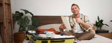 Neşeli genç bir adam, kontrol listesi olan bir turist, notlarını defterde gösteriyor, baş parmaklarını kaldırıyor, bavullarını topluyor, tatile çıkıyor, yurt dışına çıkıyor ve valizlerini hazırlıyor. Boşluğu kopyala