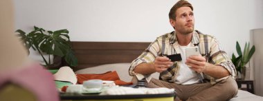 Biletleri ayırtan genç adamın portresi, tatile gitmek, kredi kartı ve akıllı telefon tutmak, giysilerle dolu çantanın yanında oturmak, tatile çıkmaya hazır olmak..