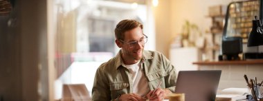 Modern bir adamın gözlüklü portresi, boş bir kafede dizüstü bilgisayarla oturuyor, not defterine notlar yazıyor, internetten okuyor, uzaktan öğreniyor, web kursundan geçiyor..