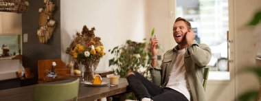 Kafede oturan, kablosuz kulaklıklı bir arkadaşla konuşan, gülen ve gülümseyen, komik podcast dinleyen yakışıklı bir gencin hayat tarzı portresi..