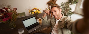 Yakışıklı ve kendinden emin bir adamın portresi selfie çekiyor, kaslarını gösteriyor, laptopun önünde oturuyor, kameraya gülümsüyor..