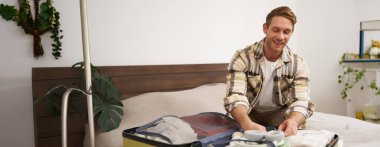 Genç bir adamın portresi, yurtdışına seyahat eden bir turist, bavula giysiler koyuyor, bavulunu kapatıyor, yatağa oturuyor, tatile hazırlanıyor. Turizm konsepti