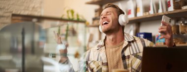 Yakışıklı mutlu adamın portresi, serbest çalışan ve kablosuz kulaklıkla müzik dinleyen, kafede sandalyesinde dans eden, kulaklıkla en sevdiği şarkıyı dinleyen..