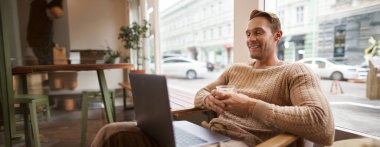 Yakışıklı genç adamın portresi kafede oturur, kahve içer ve dizüstü bilgisayarda video izler, mutlu bir gülümsemeyle ekrana bakar, çalışma alanında dinlenir..
