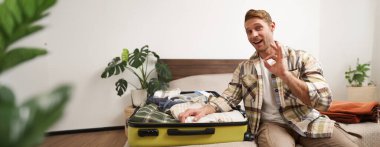 Hevesli genç adam, turist tatil için hazırlanır, bavulla yatağa oturur ve onayını gösterir, iyi kalite bagaj önerir..