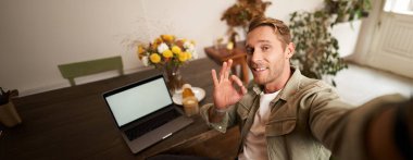 Genç yakışıklı adamın portresi, kafeterya ziyaretçisinin akıllı telefon uygulamasında selfie çekmesi, iyi referans işareti, blog yazarının kahve fiyatları, onaylı kafa sallaması..