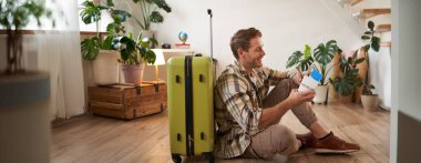 Gülümseyen yakışıklı adamın portresi, bavuluyla yerde oturuyor, elinde pasaport ve uçak biletleri var, turneye çıkıyor..