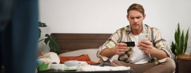 Kızgın adamın elinde kredi kartı ve cep telefonu var. Giysilerin yanında bavulda oturuyor. İnternetten ödeme yapmada sorun yaşıyor..