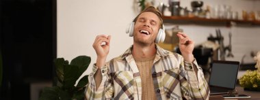 Yakışıklı, gülümseyen genç bir adam, dijital göçebe, açık alanda birlikte çalışıyor, kafeteryada çalışıyor, kablosuz kulaklıkla müzik dinliyor ve şarkı söylüyor..