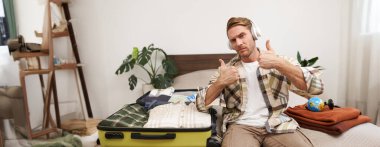 Neşeli yakışıklı bir adamın portresi, tatile çıkan bir turist, yurtdışına tatile çıkmak için kıyafetlerle bavul hazırlarken, kulaklıkla müzik dinleyen ve dans eden bir turist..