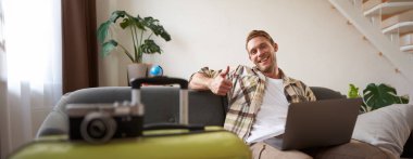 Dizüstü bilgisayarı olan neşeli bir adam, turist valizin yanına oturur, başparmağını kaldırır, tatil için yer ayırtır, tatile gider. Turizm konsepti