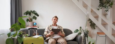 Yurtdışına giden genç bir adamın dikey fotoğrafı, tatil için bavul hazırlama, dijital tablet tutma, oteli internetten ayırtma. Turizm ve seyahat acentası kavramı