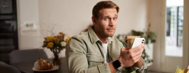 Kafedeki genç adamın portresi, elinde bir fincan kahve ve cep telefonu, kuşkucu bir yüzle kameraya bakıyor ve sırıtıyor..