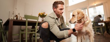 Evcil hayvan dostu kafede köpeğiyle vakit geçiren, Golden Retriever 'ı okşayan, eğlenen, ona ikramda bulunan mutlu adamın portresi..