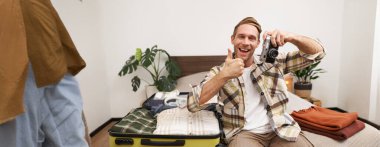 Yakışıklı turist portresi, kamera tutan erkek manken, açık bavulun yanında elbiseli adam tatile çıkıyor, yaz tatili için hazırlanıyor..