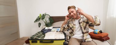 Sarışın, kulaklıklı yakışıklı bir adam, bavul ve kıyafetlerle yatakta oturuyor, tatil için hazırlanırken kablosuz kulaklıkla müzik dinliyor, seyahate çıkıyor..