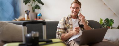 Kredi kartı tutan ve dizüstü bilgisayar kullanan bir adamın resmi. Tatil için bavul ve kamera hazırlamış. Turizm konsepti