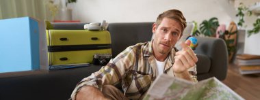 Yol haritası olan yakışıklı bir adam, kalp hareketi gösteriyor, oturma odasında bavuluyla oturuyor, tatil planları yapıyor..