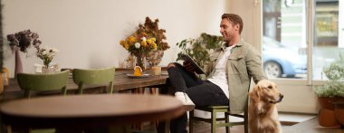 Yakışıklı genç bir adamın portresi, kafe ziyaretçisi, köpeğiyle oturuyor Golden Retriever 'ı okşuyor ve dijital tablet, yaşam tarzı ve evcil hayvan dostu kamusal alanlar hakkında haberler okuyor..