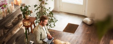 Yakışıklı iş adamının dikey görüntüsü, dizüstü bilgisayarlı genç adam masaya eğiliyor, köpeğiyle birlikte çalışma alanında oturuyor. Genç satıcı mesajlarını kontrol ediyor, gülümsüyor, bir ofiste evcil bir hayvanla çalışıyor..