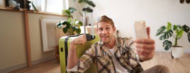 Komik yakışıklı adam, turist kasları kasıyor, tatile çıkıyor, bavulla oturuyor, akıllı telefondan selfie çekiyor. Turizm ve seyahat kavramı