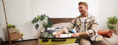 Yaz tatiline hazırlanan neşeli genç adamın portresi, tatile gitmek, bavulunu elbiseleriyle hazırlamak ve heyecanla gülümsemek. Yaşam tarzı, insanlar ve seyahat konsepti