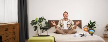 Genç turistin portresi, yatağında meditasyon yapan, yoga zen pozu veren rahat gülümseyen adam, dünya ve kamera, bavulunu hazırladı ve rahatladı, tatile çıkıyor. Farkındalık ve turizm