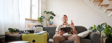 Genç adam bir şey umuyor, dijital tabletli turist, çapraz parmaklar, bavulla oturuyor, tatil için dua ediyor. Seyahat konsepti