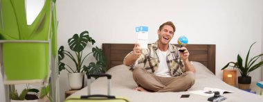 Mutlu adamın portresi, turist yurt dışına tatile çıkıyor, elinde dünya ve uçak bileti var, tatili için heyecanlı, gülümseyen, coşkulu bir yüz ifadesiyle yatakta oturuyor. Boşluğu kopyala