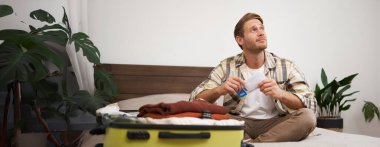 Bavullu genç bir adamın portresi, uçak biletlerini elinde tutması, tatile gitmeye hazırlanması, tatil için eşyalarını toplaması, turist kıyafetlerini çantaya koyuyor..