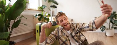 Gülümseyen yakışıklı adamın portresi fotoğrafta bisepslerini gösteriyor, akıllı telefonda selfie çekiyor, bavulun yanına oturuyor, tatil turuna çıkıyor..