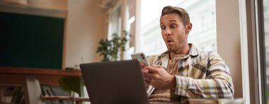 Kafedeki adamın görüntüsü, kafeterya ziyaretçisinin dizüstü bilgisayarı ve akıllı telefonuyla koltukta oturması, cep telefonu ekranına şaşırmış ve heyecanlı görünmesi, büyük haberlere hayran kalması, uygulamanın tanıtım teklifi..