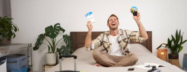 Mutlu adamın portresi, turist yurt dışına tatile çıkıyor, elinde dünya ve uçak bileti var, tatili için heyecanlı, gülümseyen, coşkulu bir yüz ifadesiyle yatakta oturuyor. Boşluğu kopyala